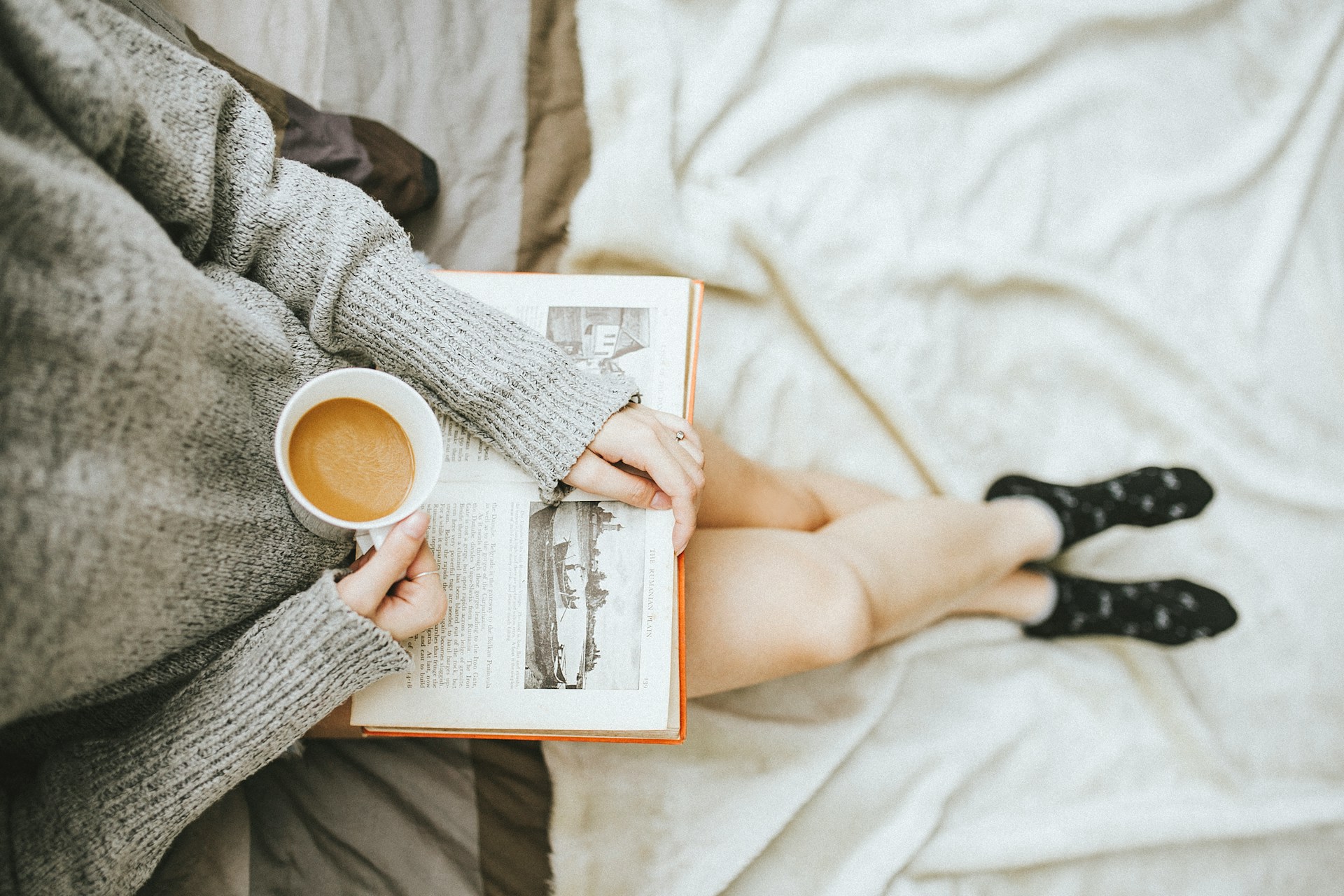 A woman reading a book in her lap and drinking a cup of coffee